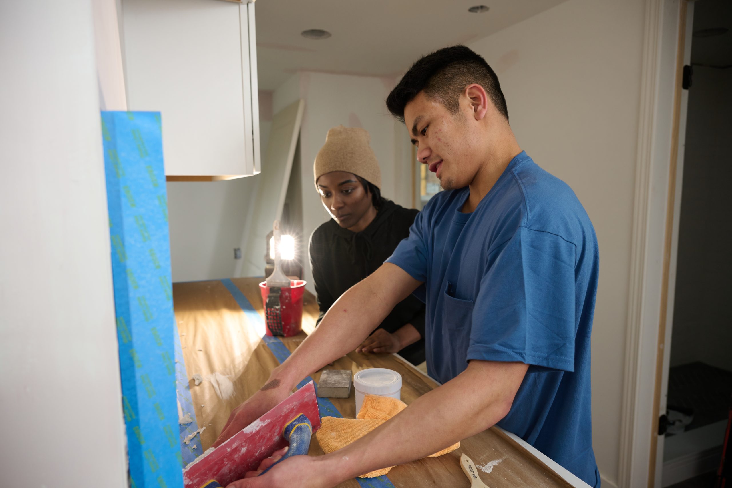 - Asian male teaching African American female how to apply grout to tile in a kitchen